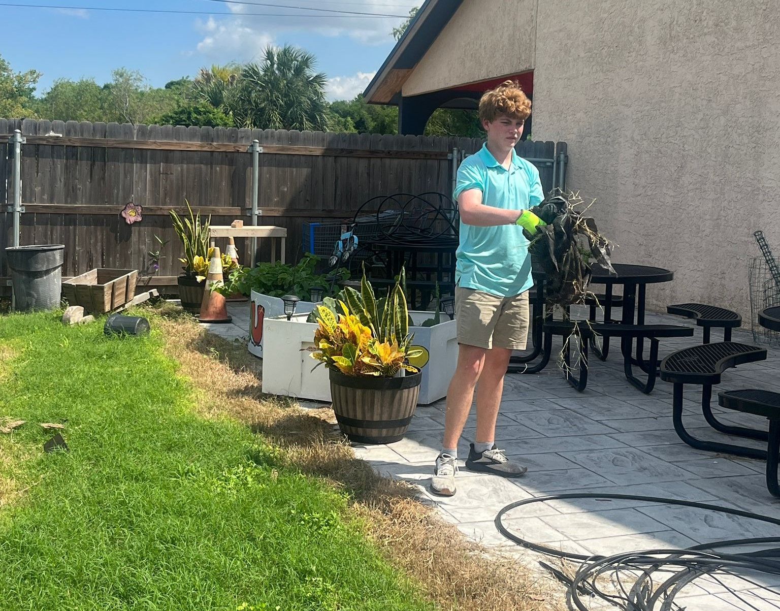 Teenager in shorts and a blue shirt holding a rope in a backyard with plants and picnic tables.