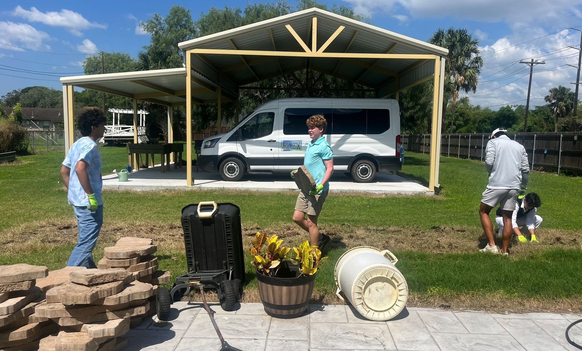 People working outdoors near a white van under a shelter. They are preparing a garden bed with tools and bricks.