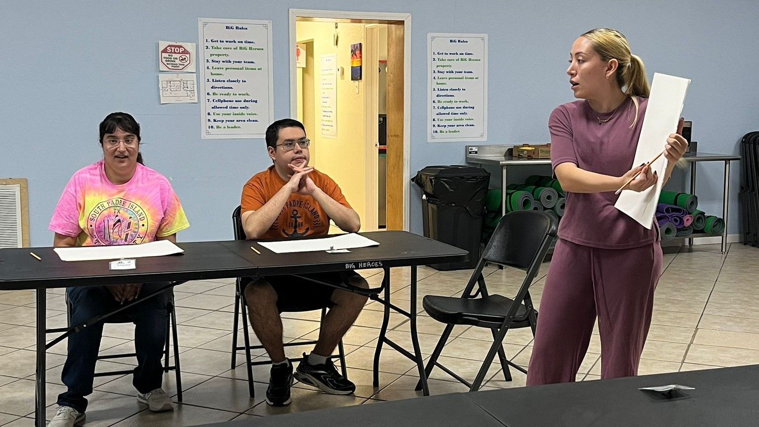 A woman is holding a piece of paper in front of a group of people sitting at tables.