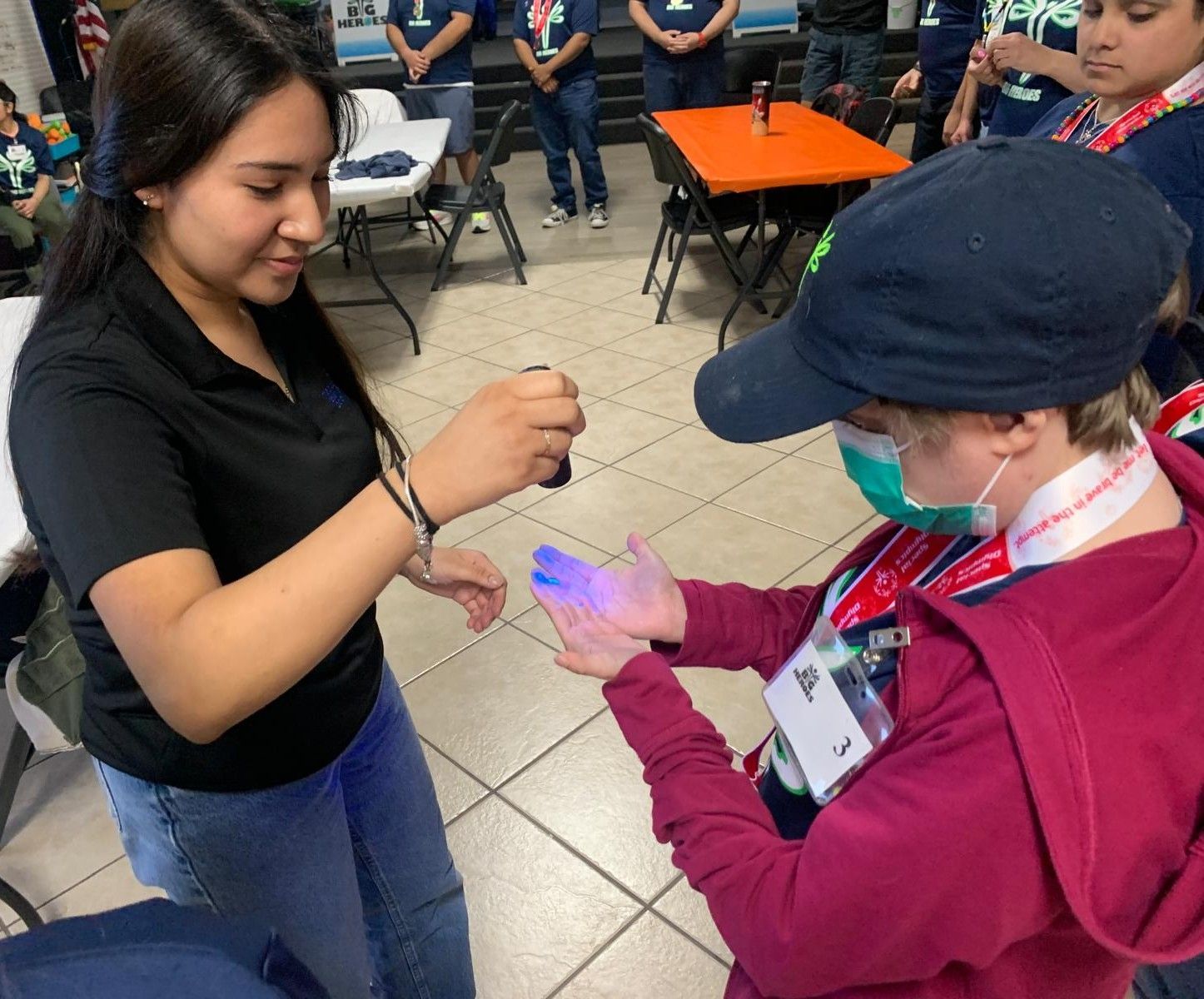 Woman shining UV light on another's hand, checking for germs. Indoors, others watch.