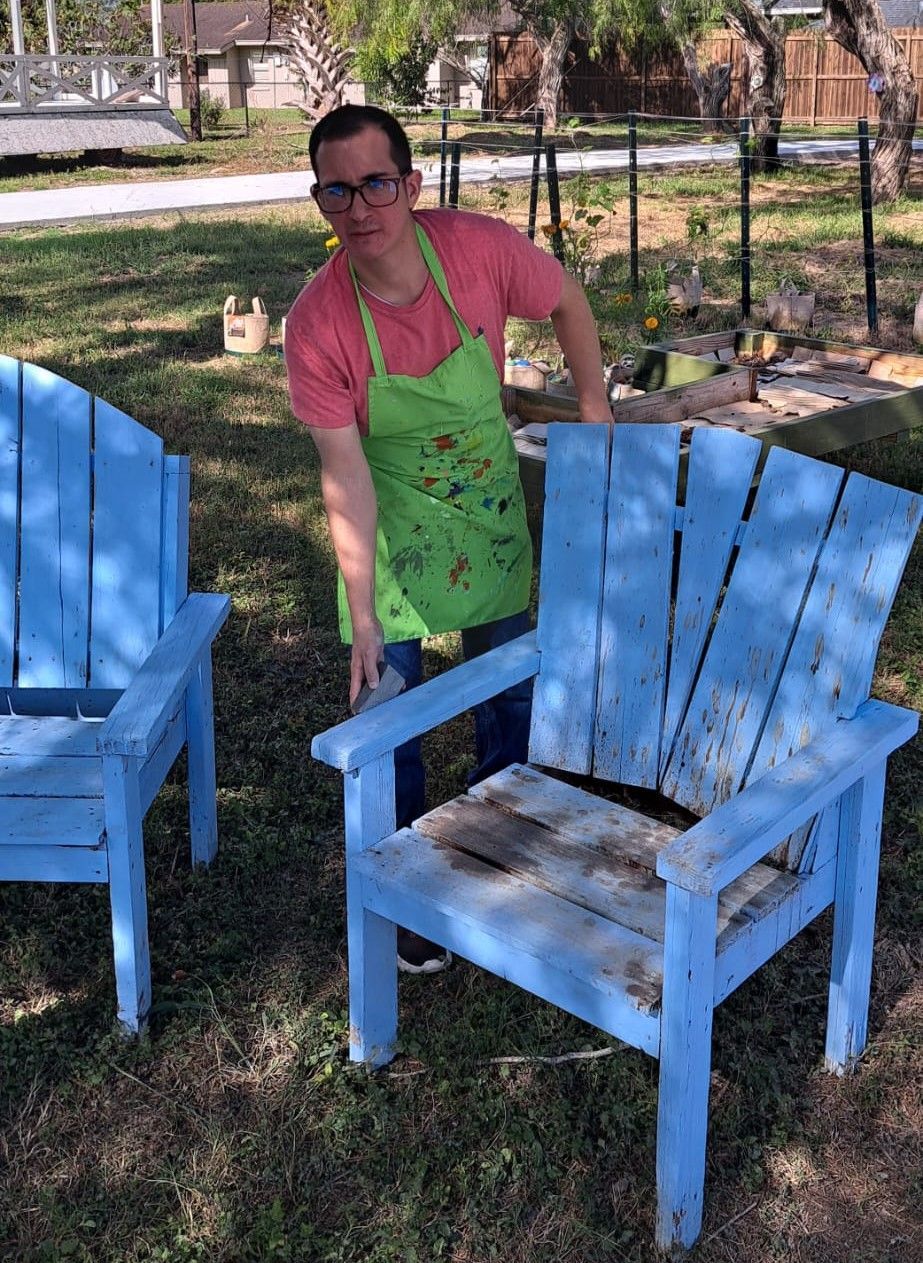 A man in an apron is standing next to two blue wooden chairs.