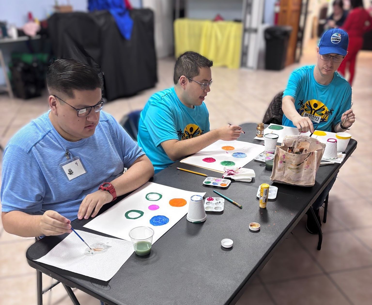 Three men are sitting at a table painting with watercolors.