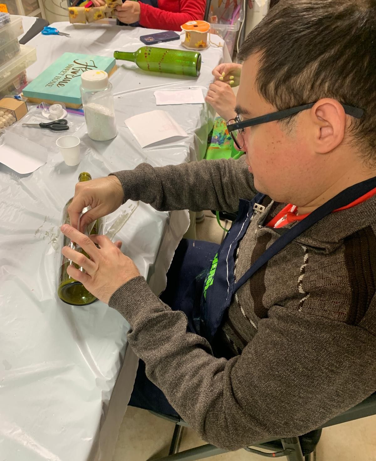 A man in a wheelchair is sitting at a table cutting a glass bottle