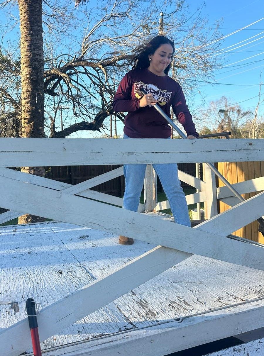 Woman in maroon sweatshirt and jeans leaning on a white fence, smiling. Outdoors, bright.