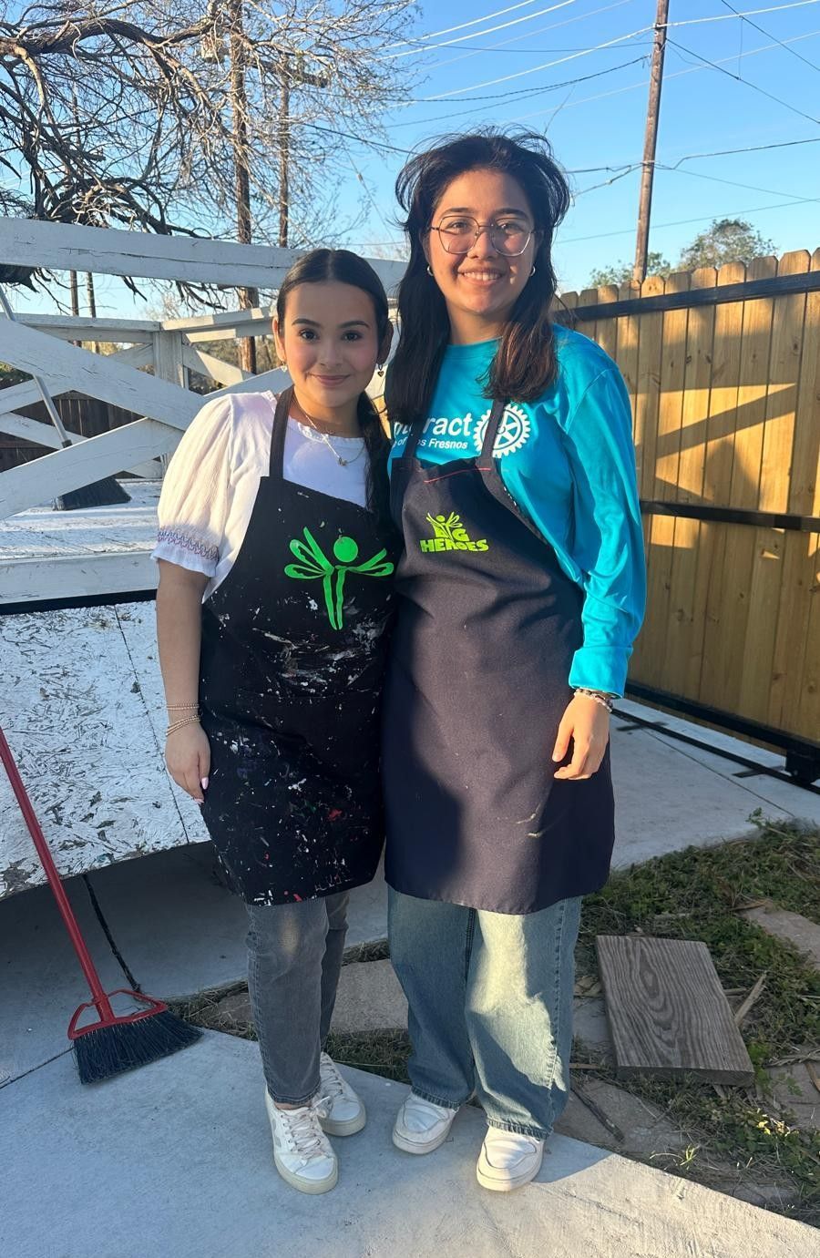 Two young women in aprons stand outside smiling, one in a blue shirt, the other in white.