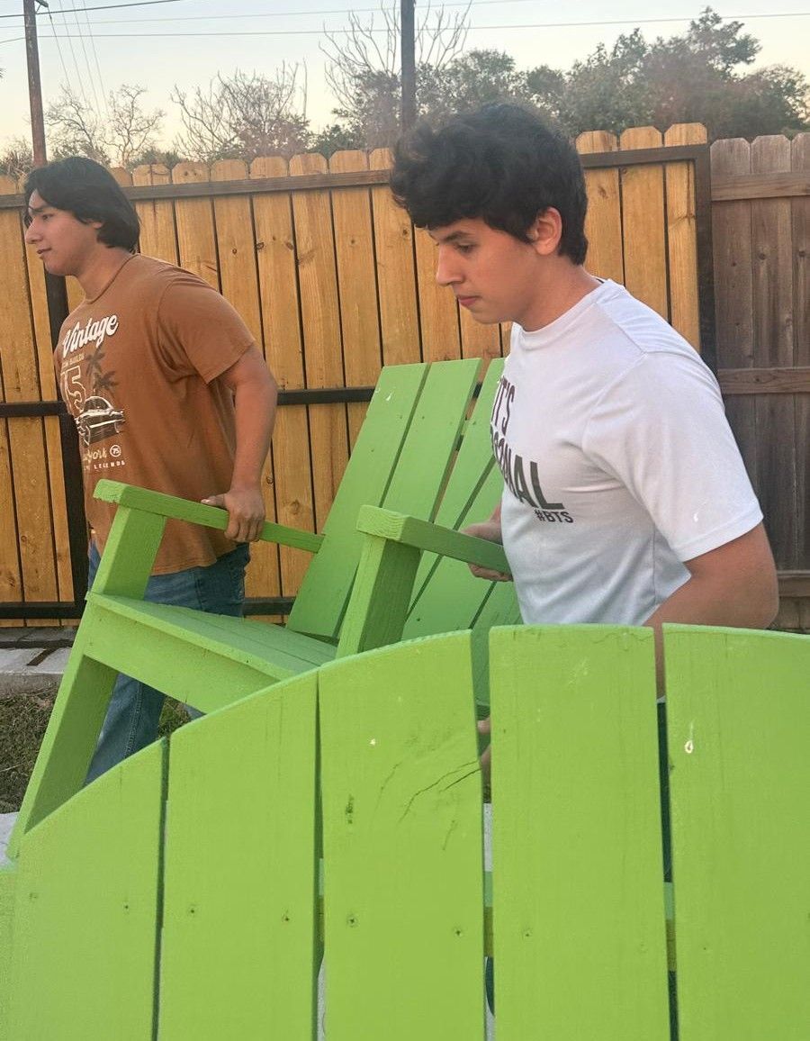 Two young men carrying a bright green Adirondack chair outside near a wooden fence.