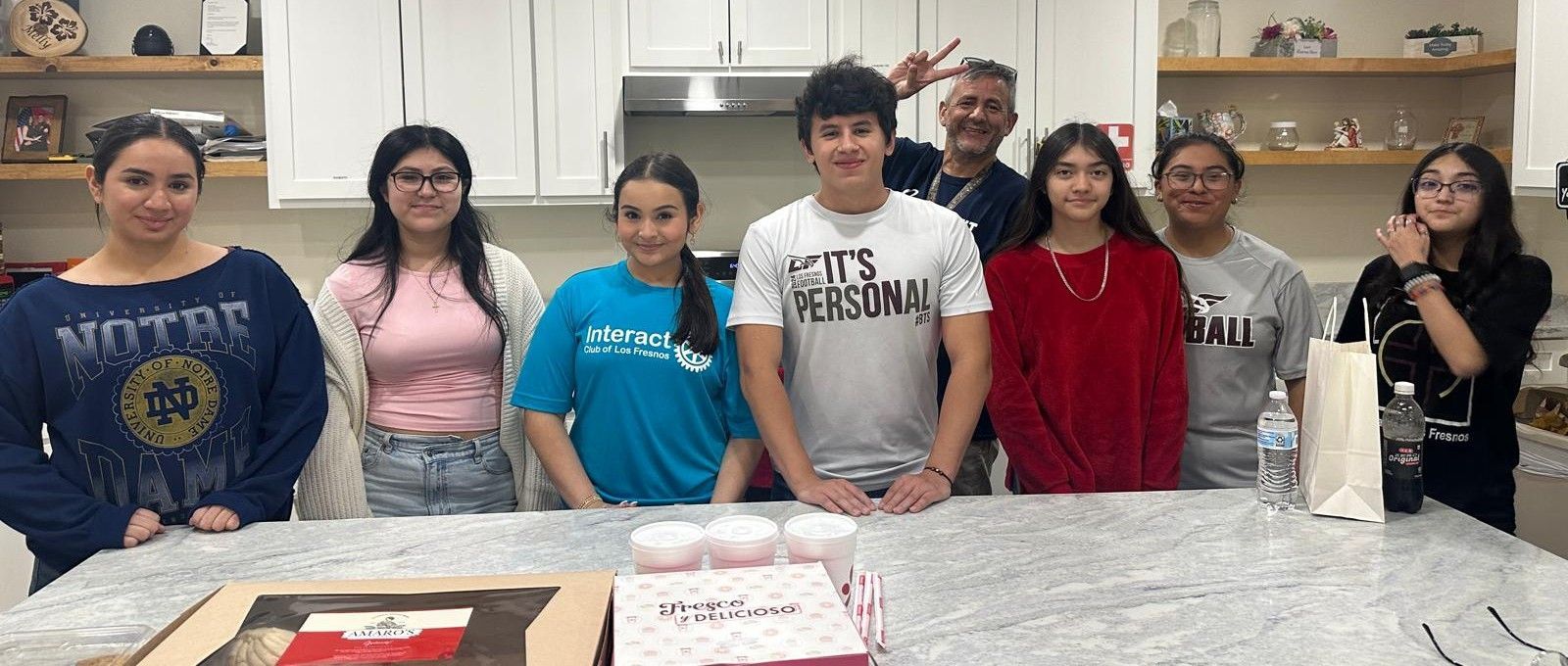 A group of people in a kitchen smiling, some are pointing, with food and cake in front of them.