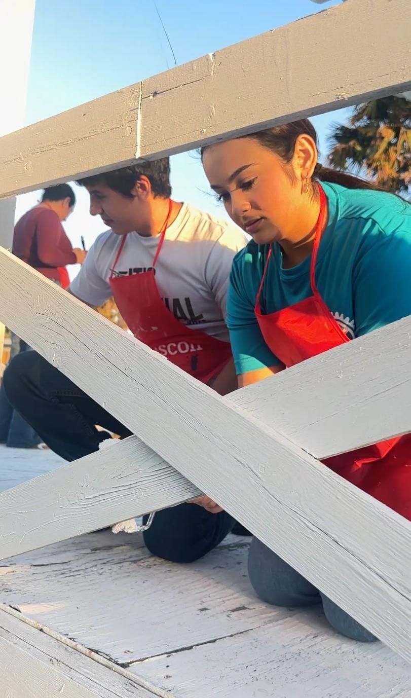 Two people, wearing aprons, painting a white fence outdoors.