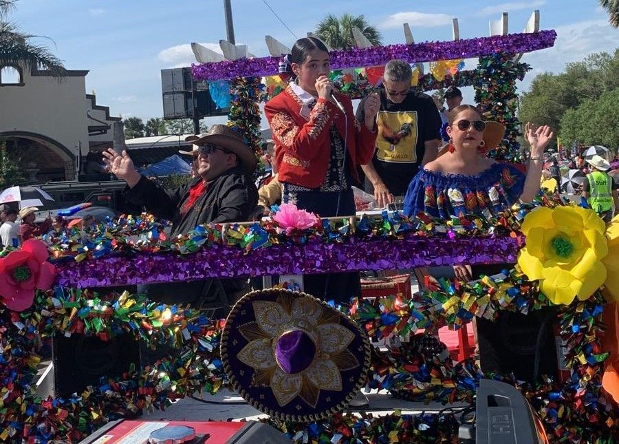 People on a decorated float in a parade, waving. Celebratory with Mexican colors and decorations.