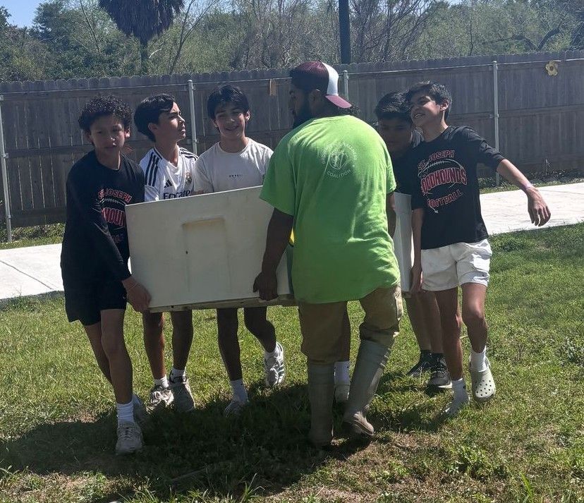A group of boys and a man carrying a large white object on grass outside; sunny day.