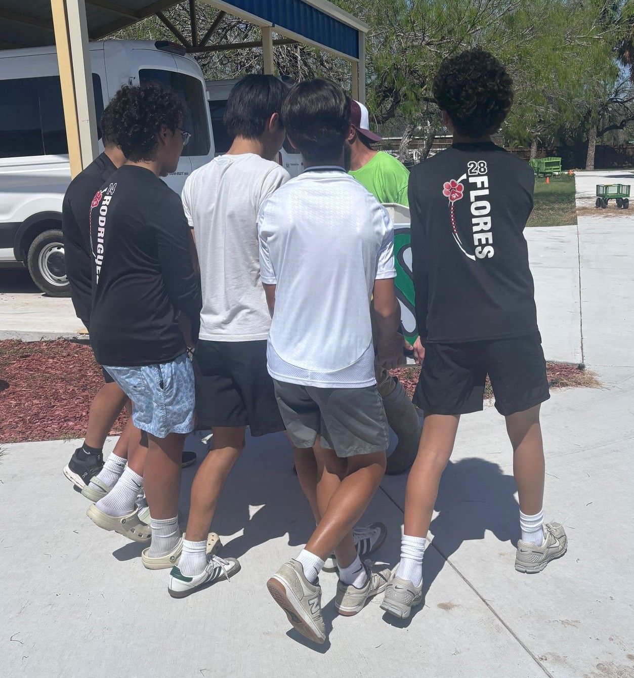 Group of young men standing in a circle on concrete; some wearing athletic clothing.