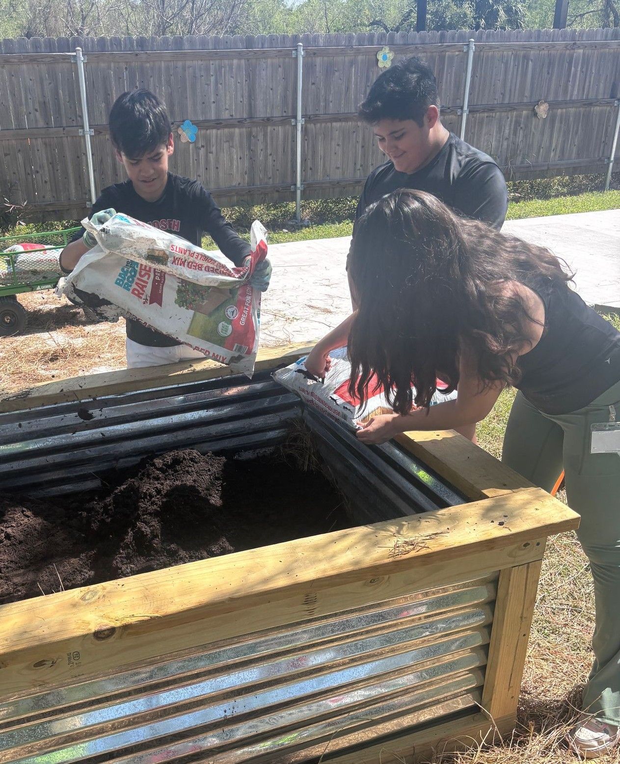 Three children filling a raised garden bed with soil outdoors.