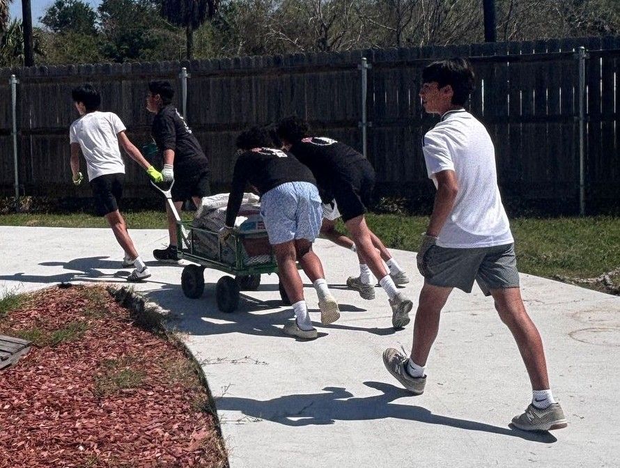 Group of teens pushing a cart loaded with bags of material on a paved walkway outdoors near a fence.
