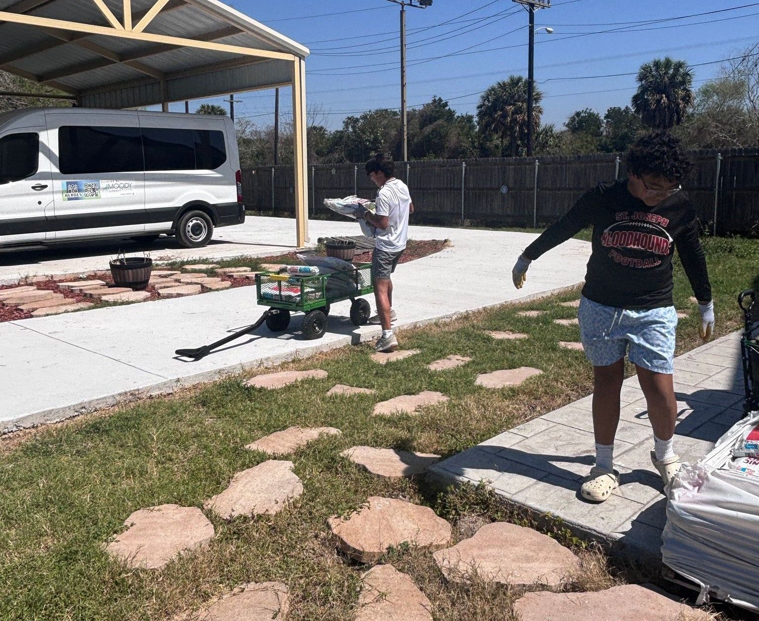 Two young men outdoors moving supplies near a white van and walkway, sunny day.