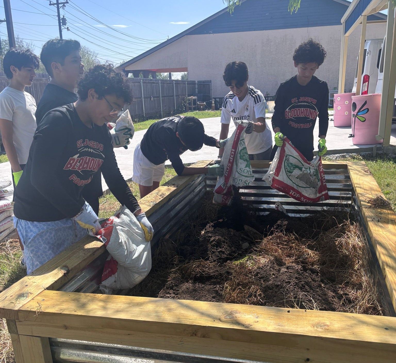 A group of boys add soil to a wooden garden bed outdoors on a sunny day.