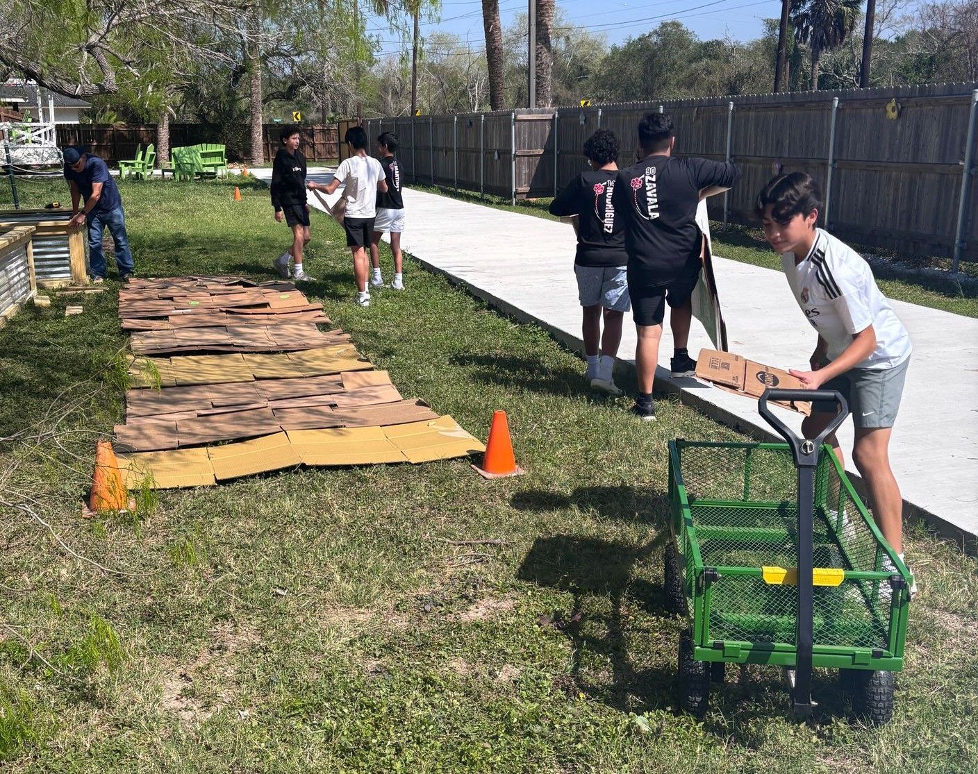 People working on a garden path; one pulls a green cart, others lay down cardboard, sunny outdoors.