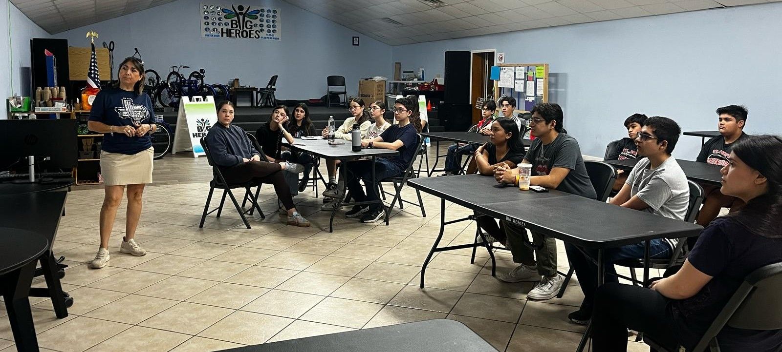 A woman presents to a group of students seated at tables in a classroom.
