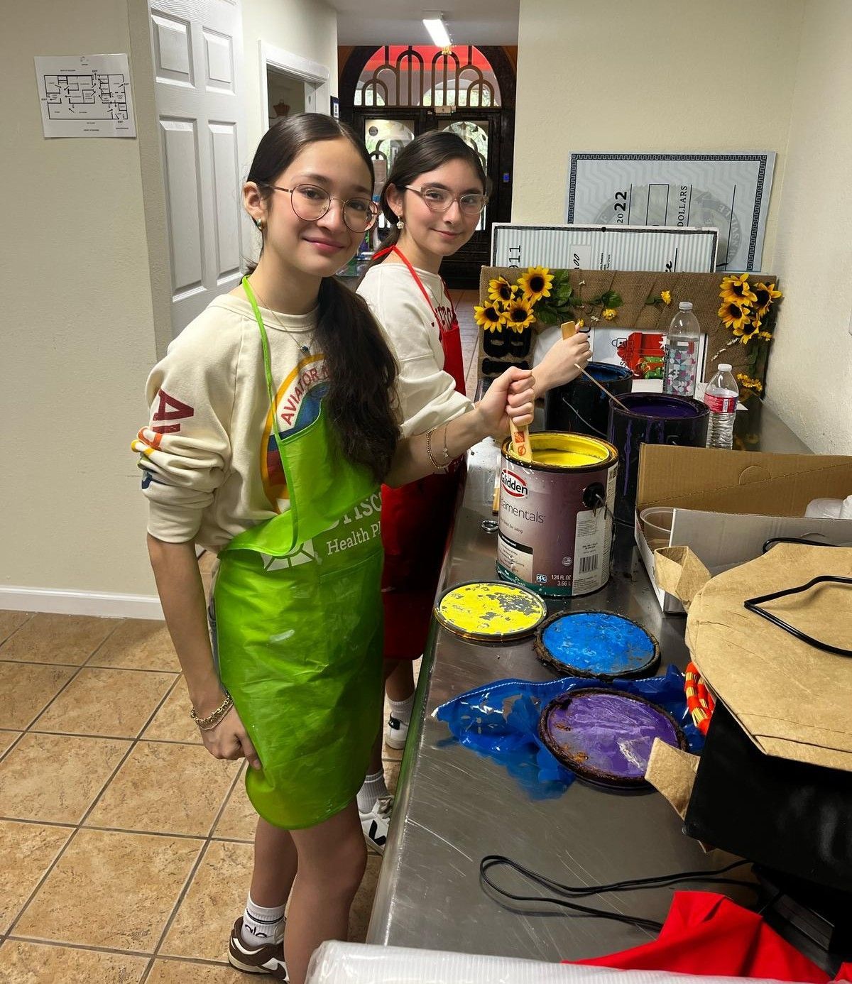Two young women with aprons prepare paint in a room. One smiles, holding a paint stick.