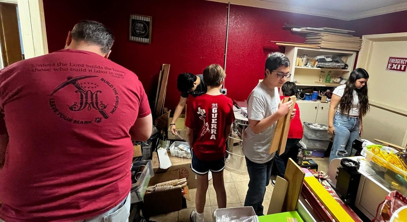 People in a cluttered workshop, some holding wooden boards, working. Red wall, various items visible.
