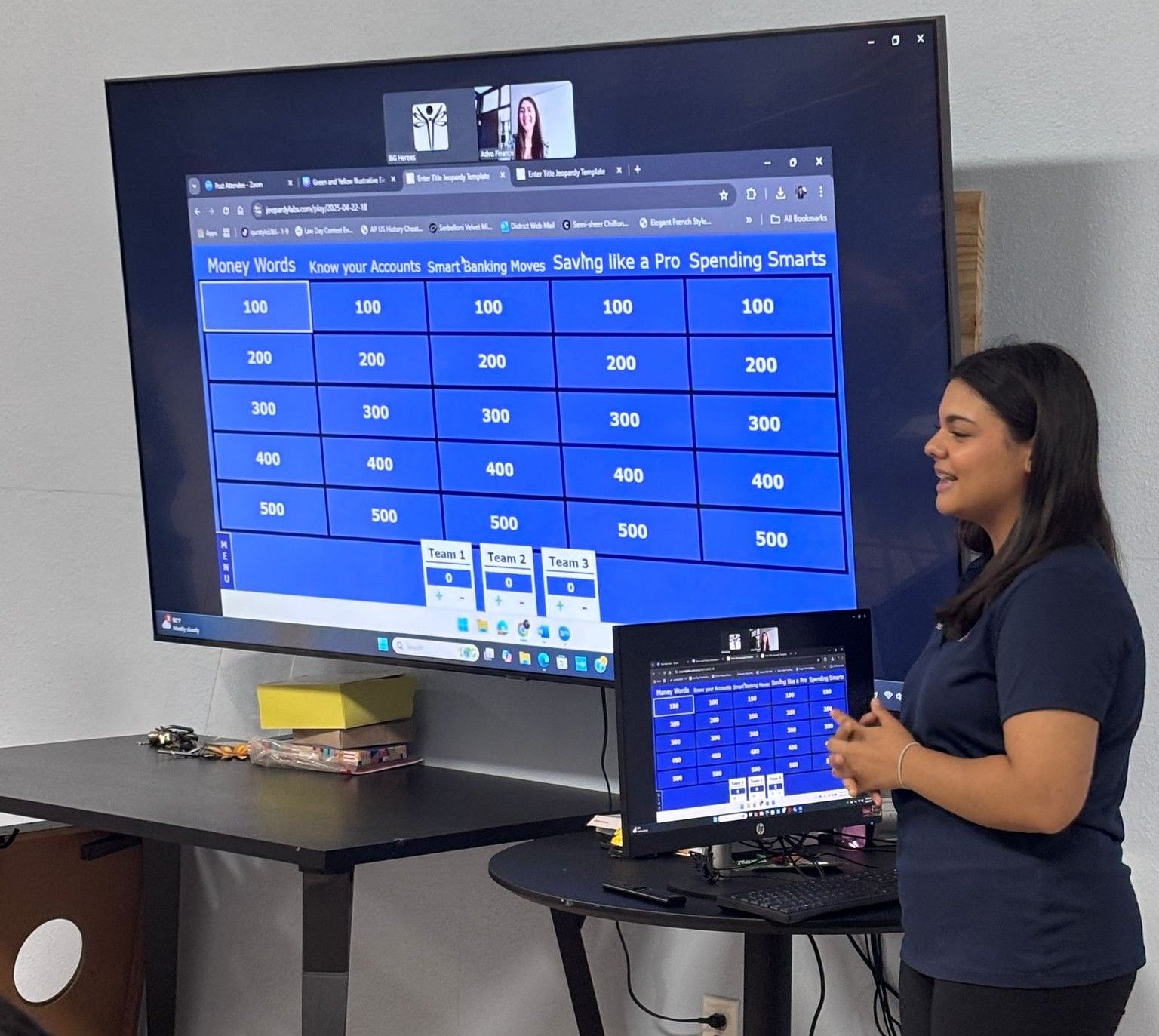 Woman presenting a Jeopardy-style game on a large screen. A smaller screen is on a table.