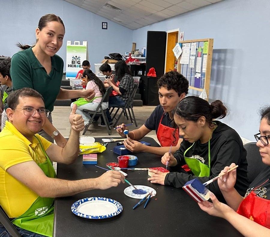 People painting together at a table; instructor smiles, gives thumbs up. Bright colors, indoor setting.