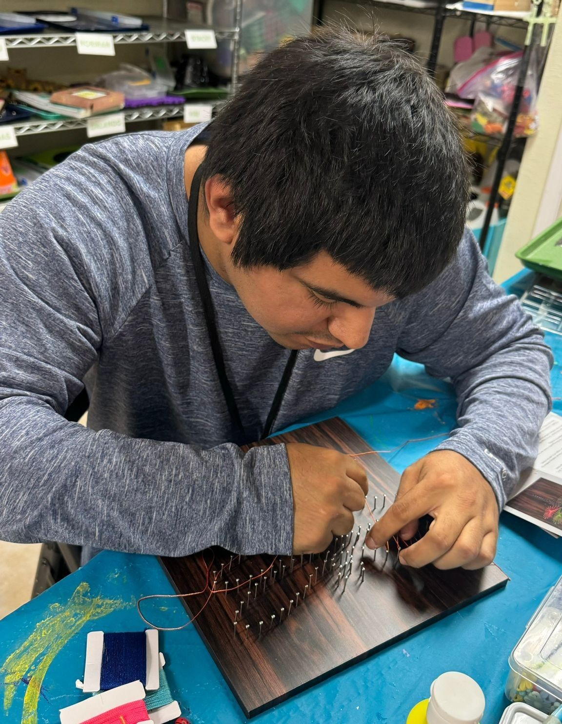 Person with dark hair crafting string art, focused, at a table, with blue and brown surfaces.