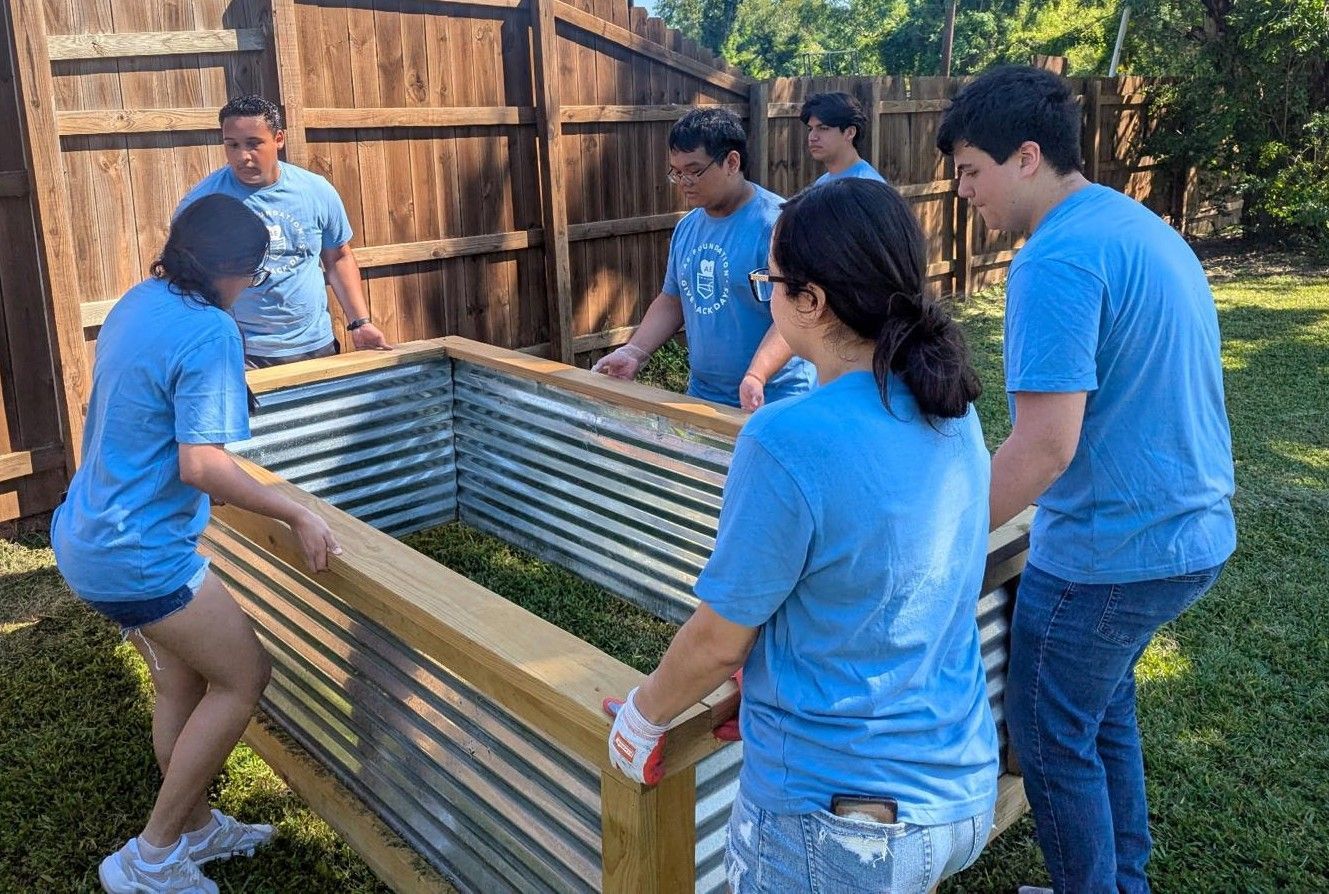Group of people in blue shirts assembling a corrugated metal and wood planter box outdoors.