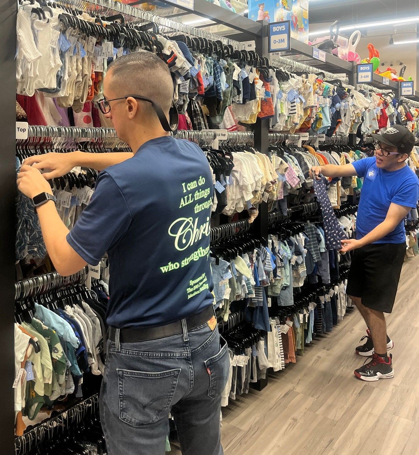 Two people browsing baby clothes in a store. One in blue shirt with text, other in glasses, both sorting.