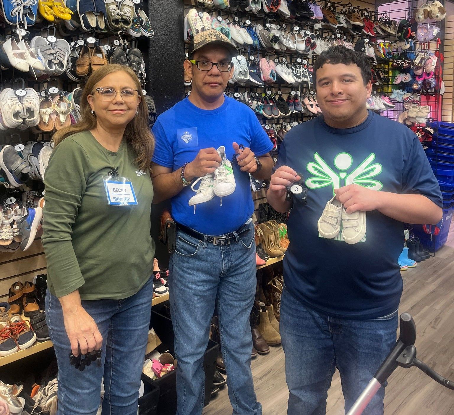3 people hold up shoes in a shoe store. The person on the left green shirt, middle a blue shirt, & right in dark blue shirt.