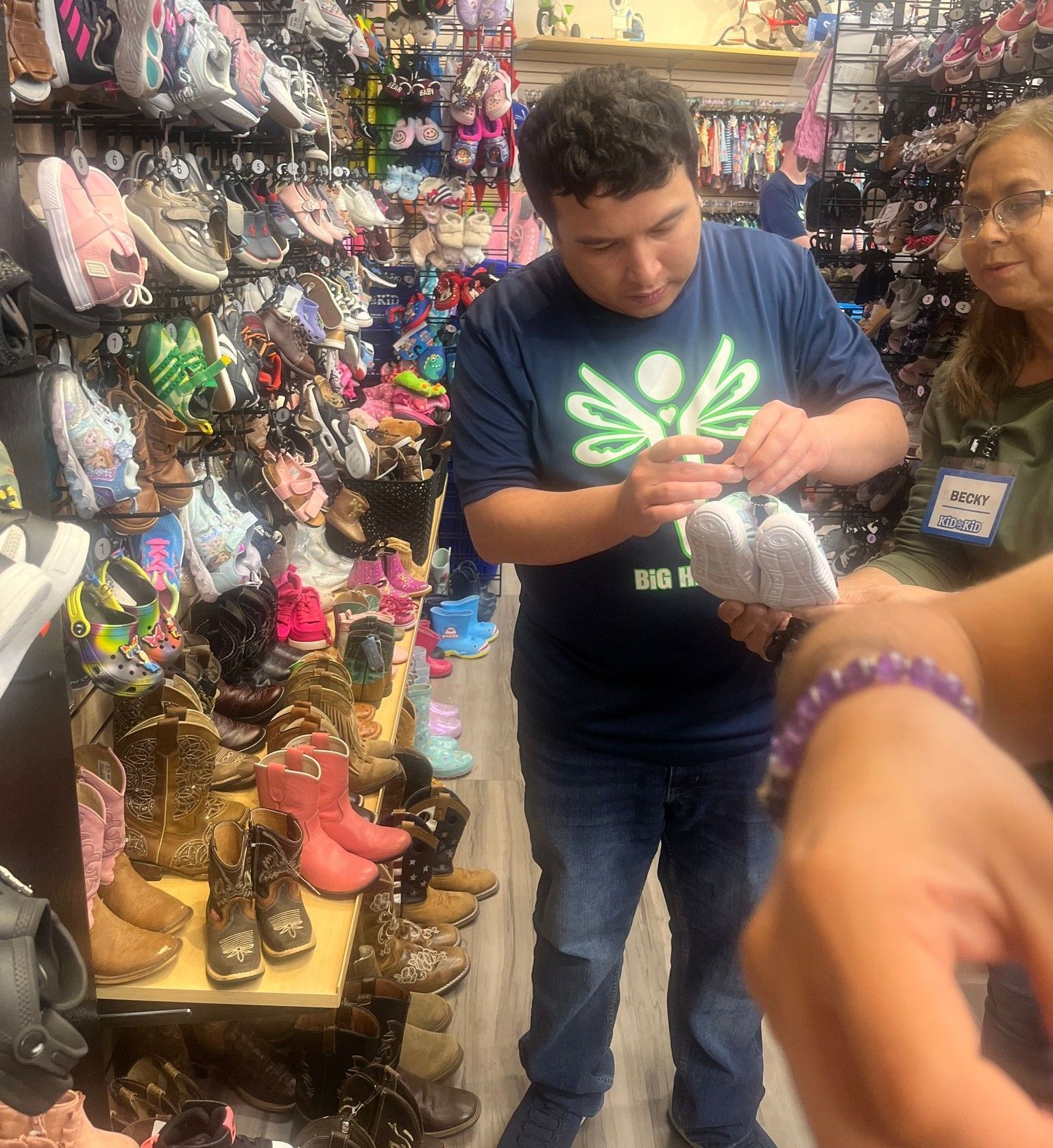 Man looking at shoes in a store, surrounded by shelves of footwear; another person stands nearby.