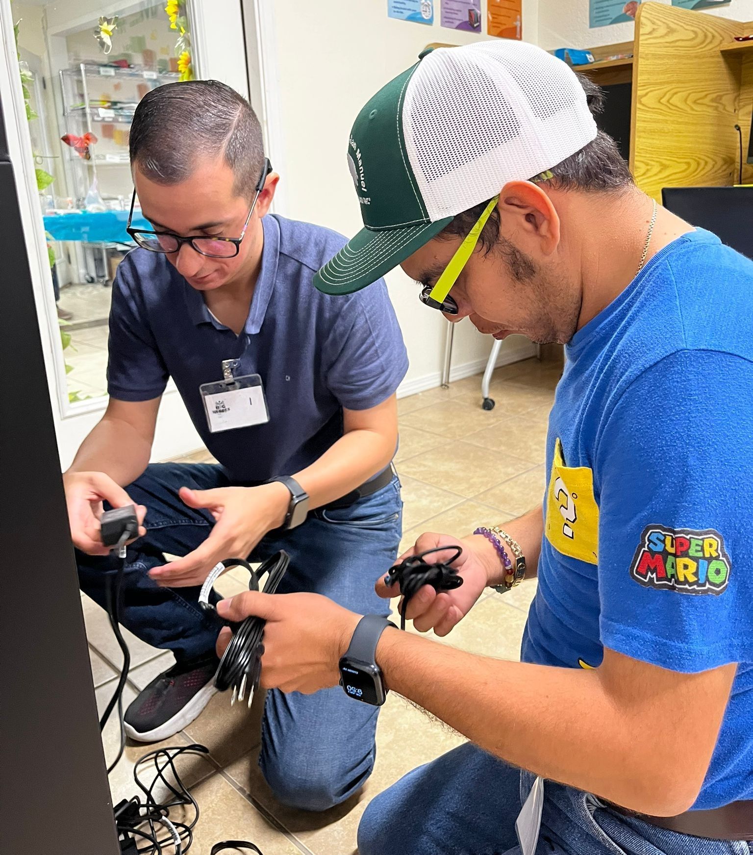 Two men connecting wires near a black panel, indoors. One wears a Super Mario shirt, the other a blue polo and glasses.