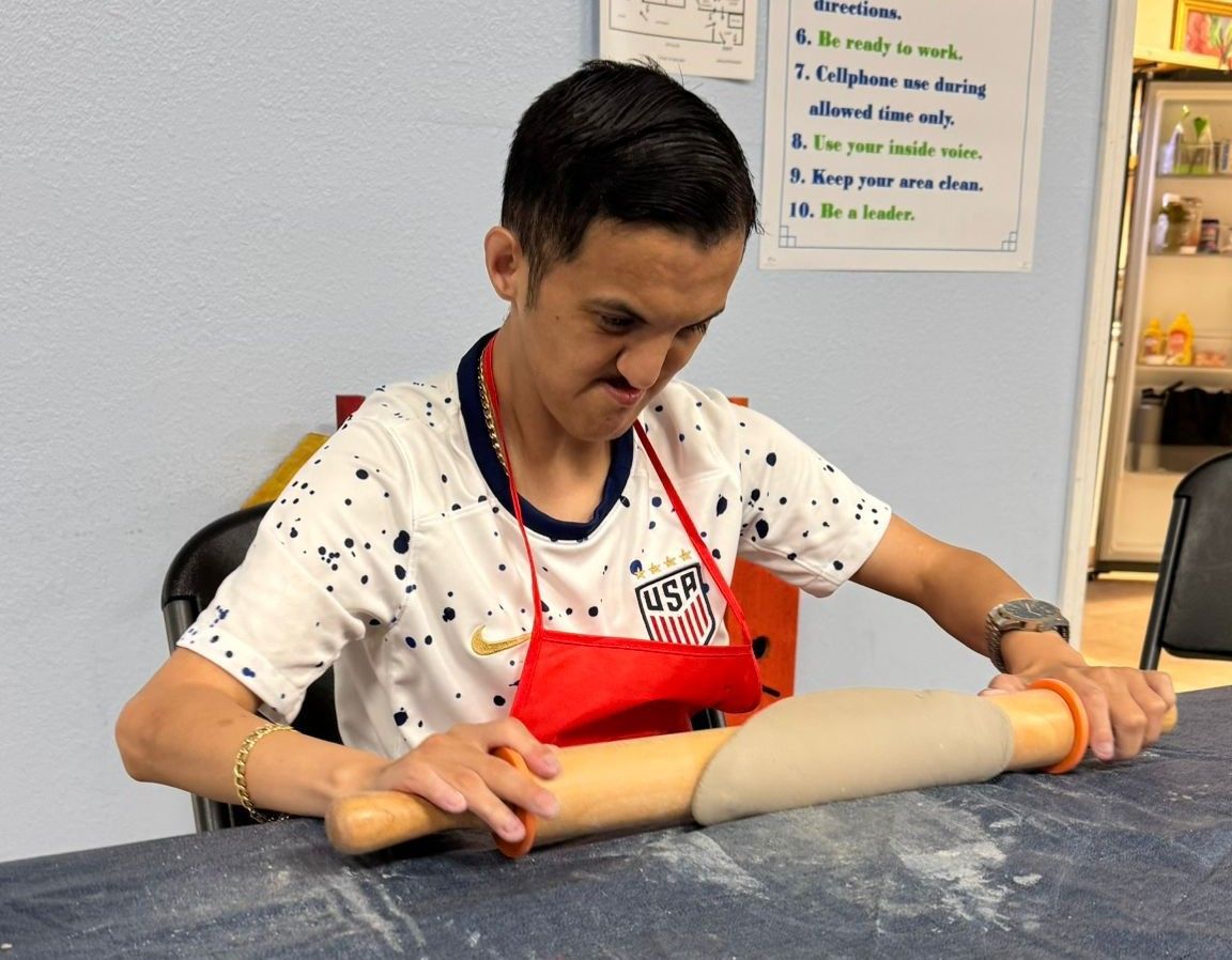 Person rolling dough with focused expression; wearing an apron in a kitchen setting.