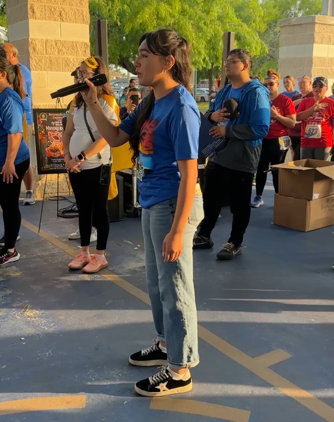 Woman with microphone speaking at an outdoor event, wearing blue shirt, jeans and sneakers. People in background.