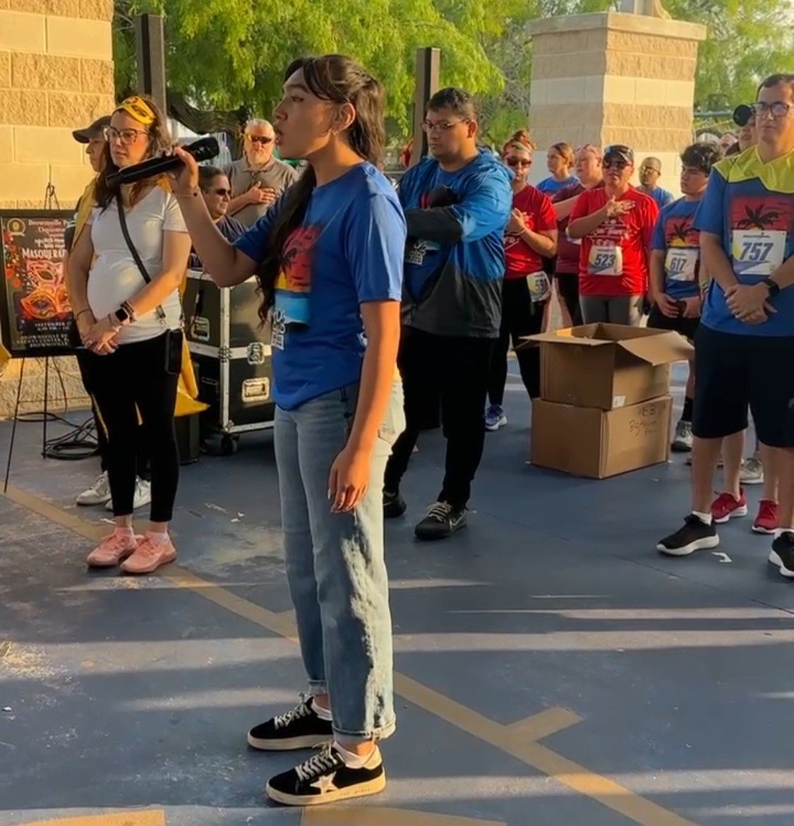 Woman speaking into a microphone at a race start. People in running gear in background, outdoor setting.