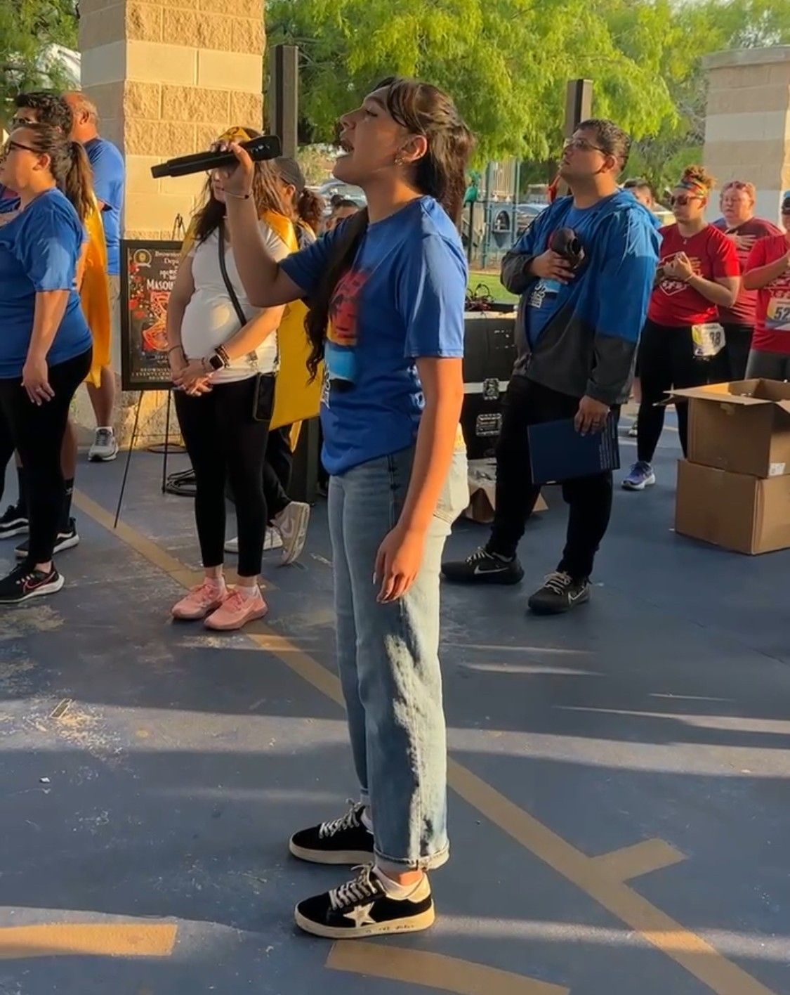 Woman singing into a microphone at an outdoor event, other people in the background, blue shirts.