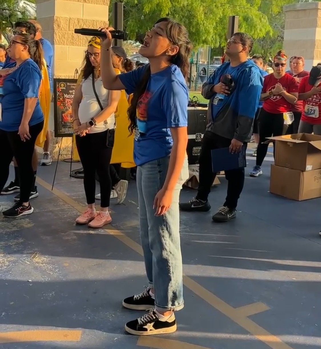 Woman singing into a microphone at an outdoor event, blue shirts, people watching.