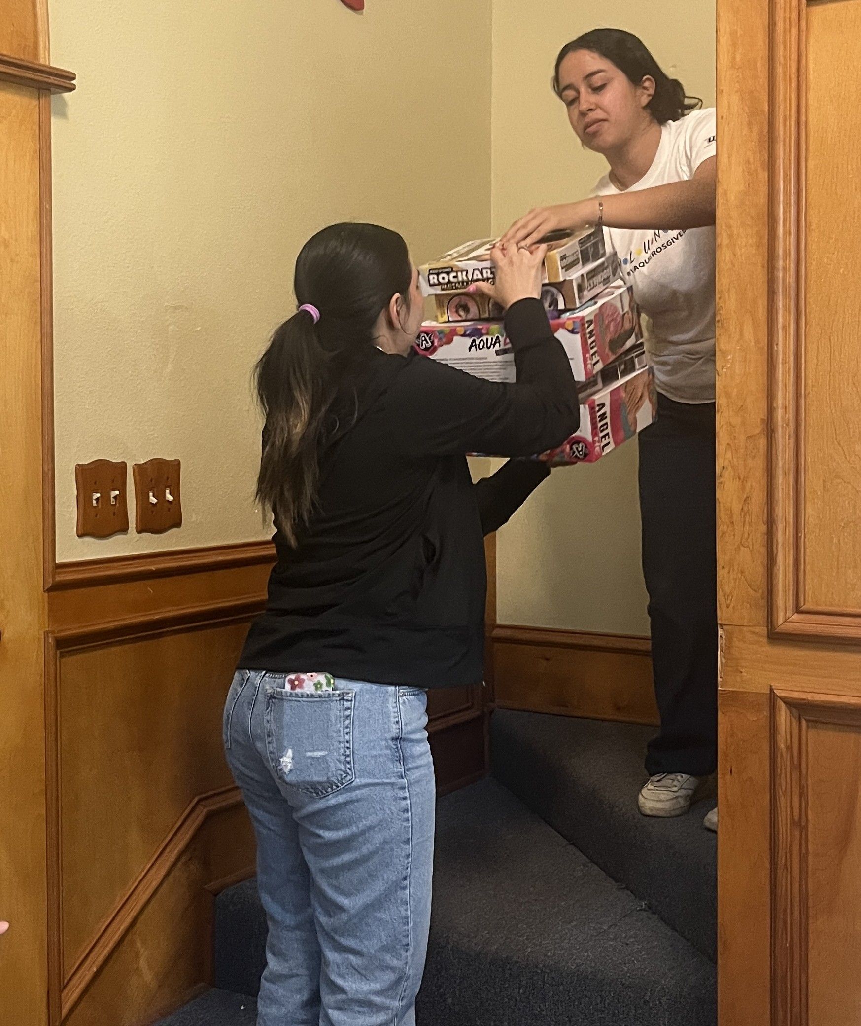 Two women carrying a large, wrapped gift up a staircase. One is in a black jacket, other in a white tee.