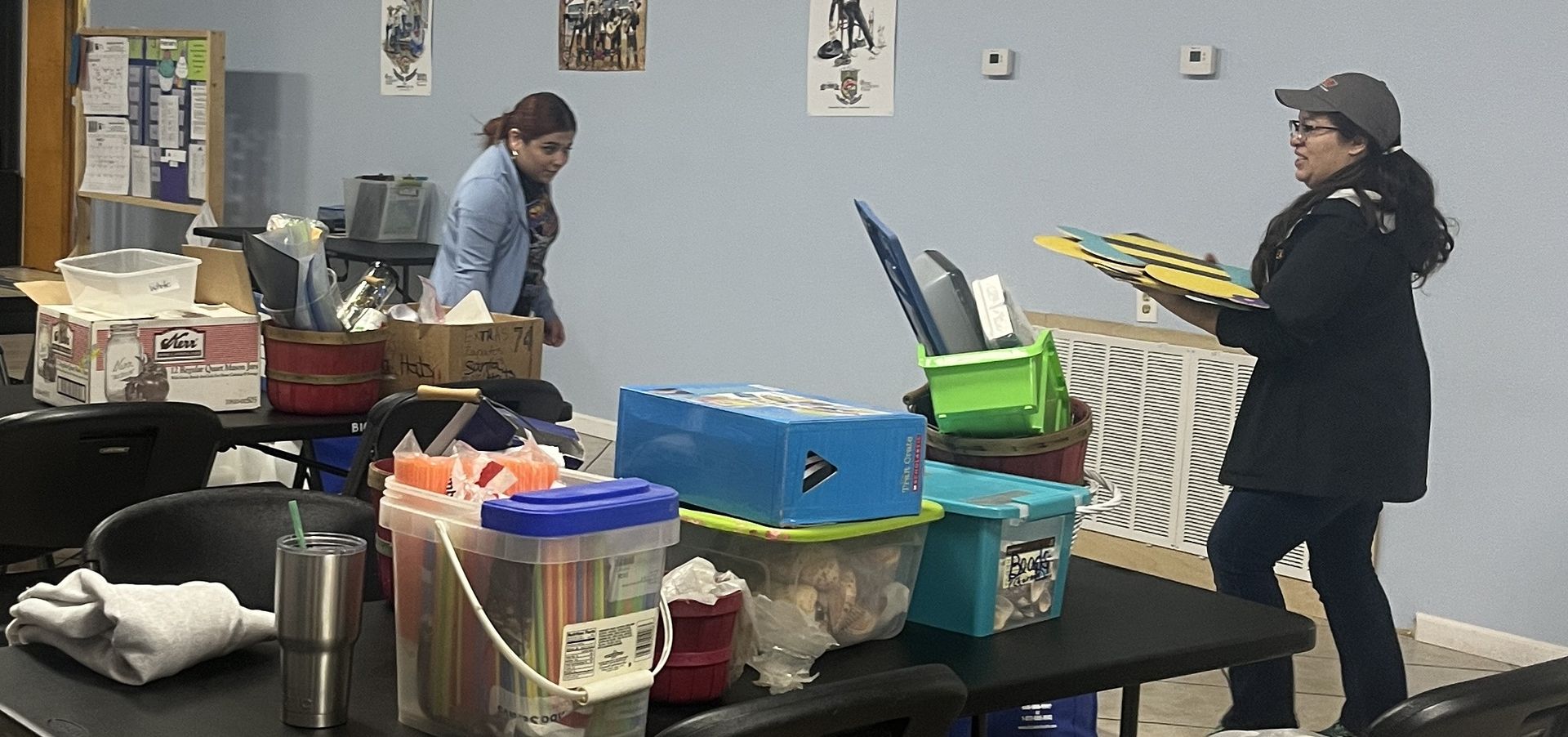 Two women organizing items in a room. One holds a board, others are on tables.