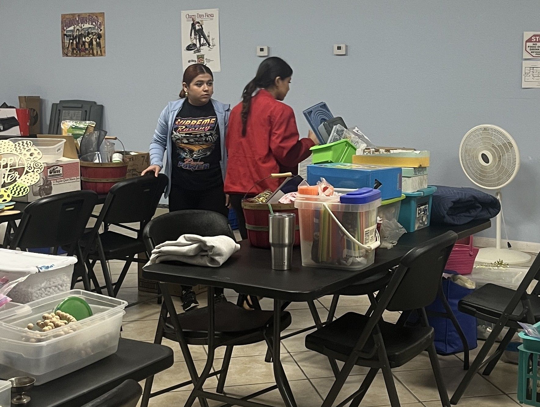 Two women standing near a table filled with items in a cluttered room.