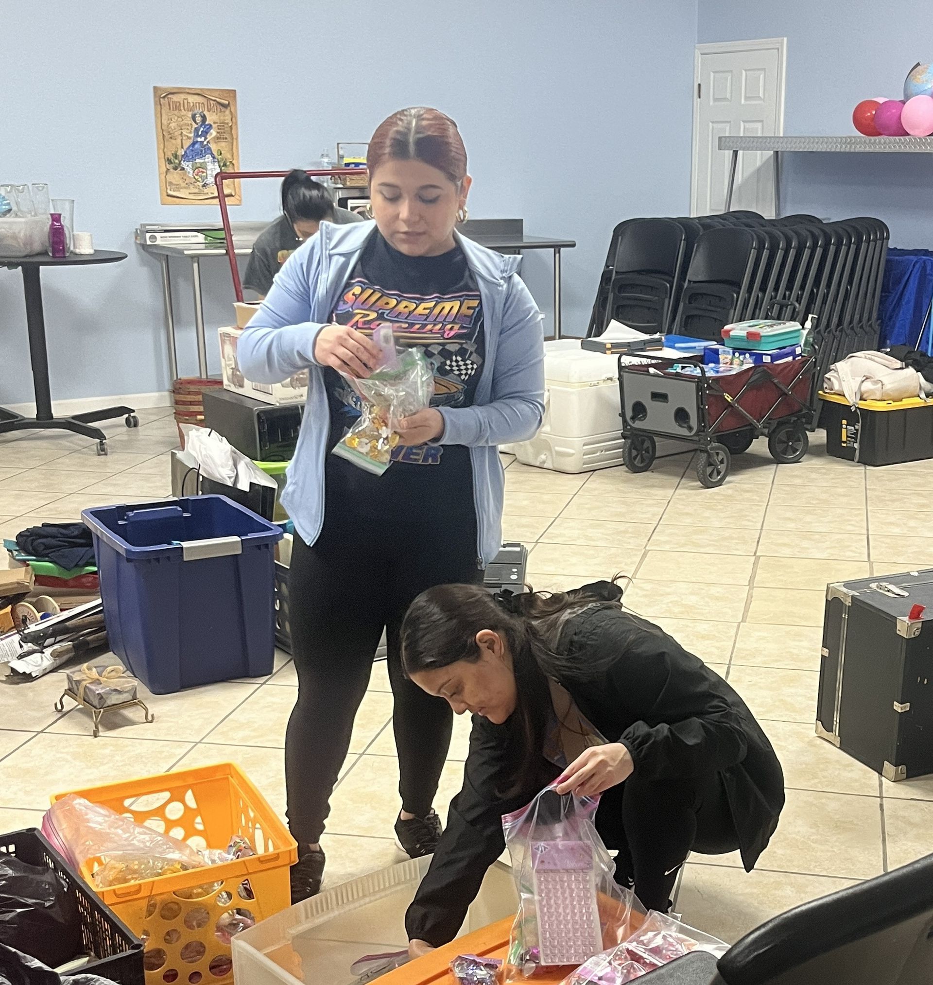 Two women packing bags inside a room. One stands, holding a bag, while the other kneels, sorting items in a bin.