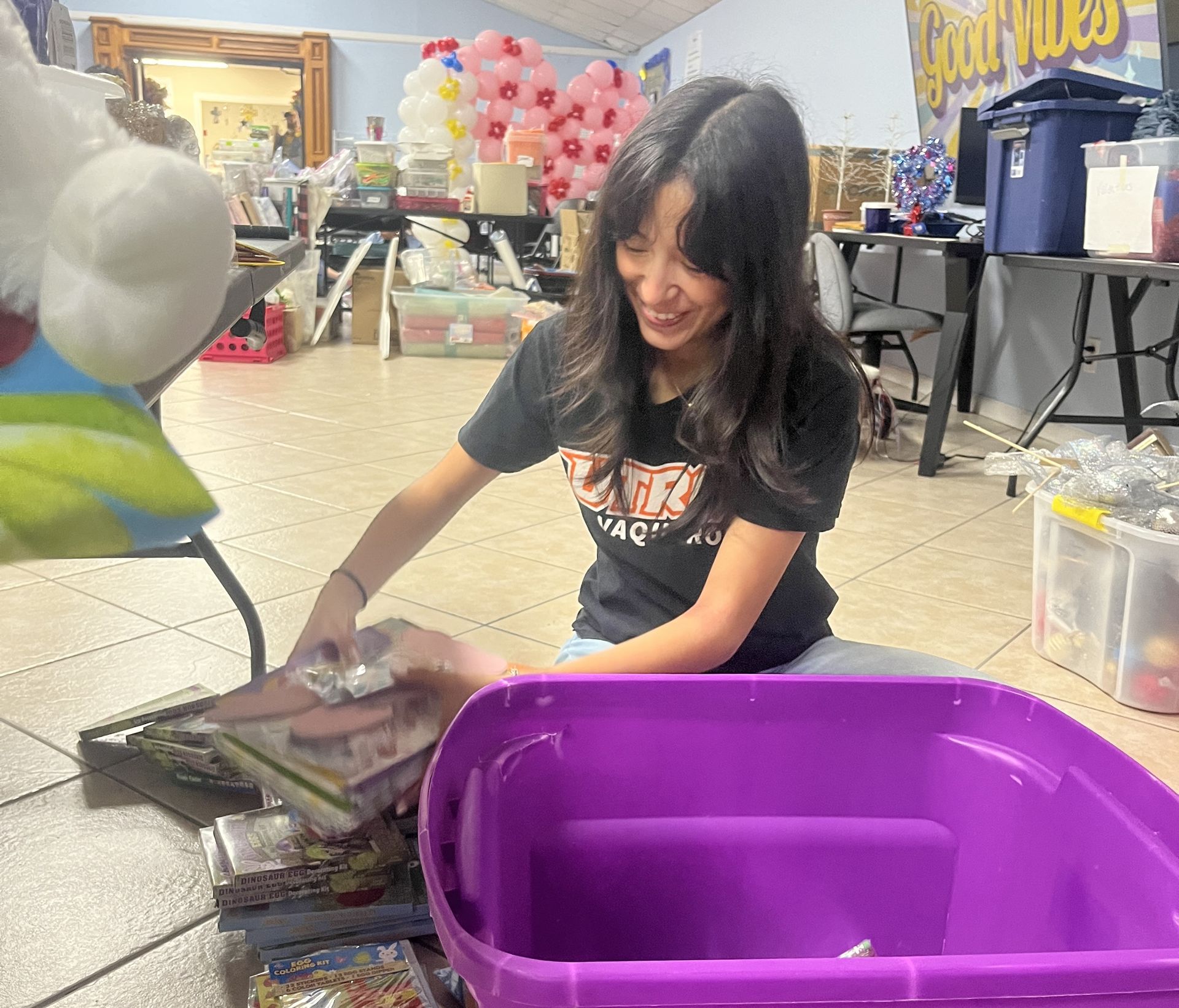 Woman smiling, organizing items near a purple bin in a room decorated with balloons and tables.