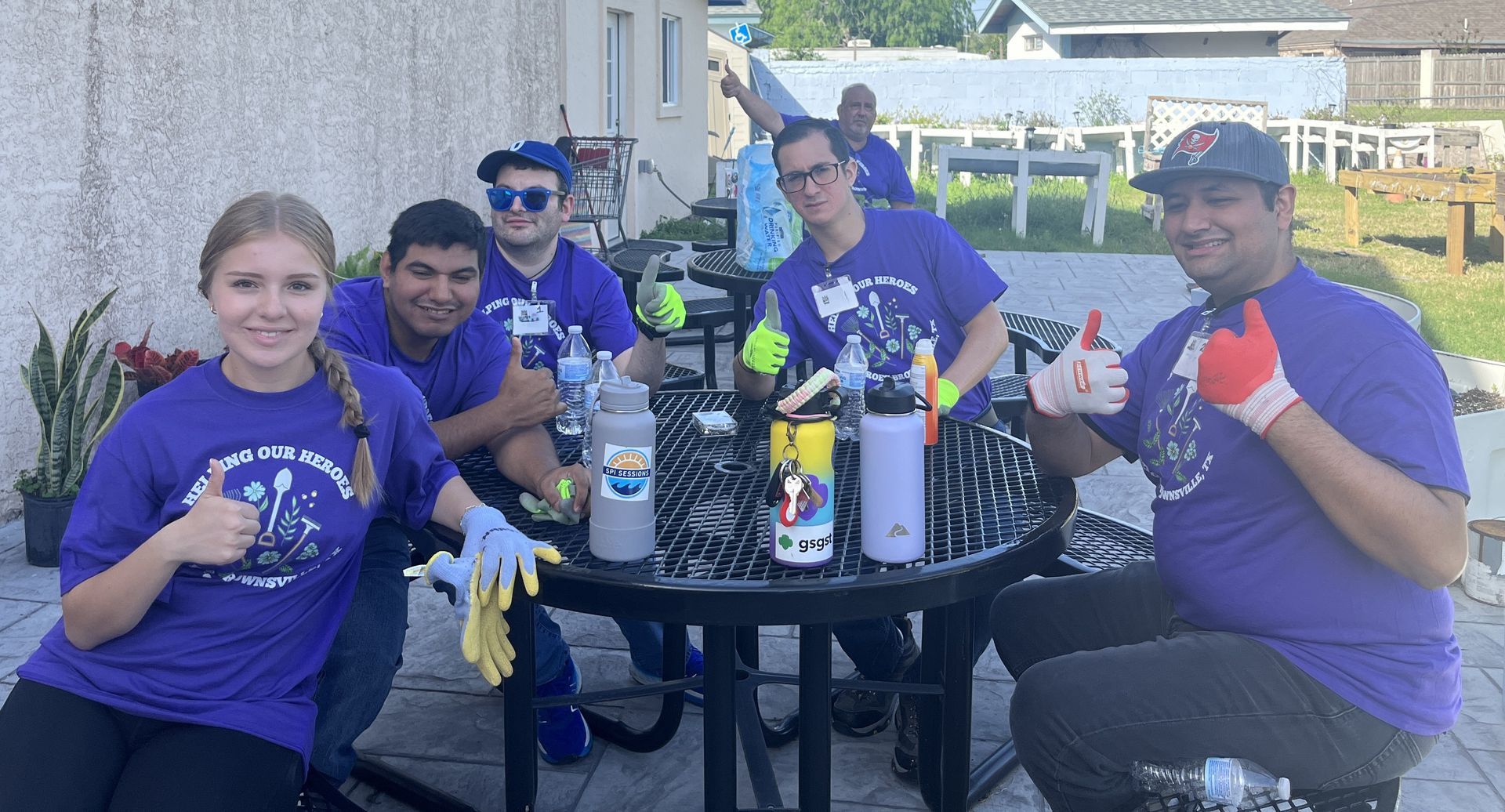 Group of people in purple shirts painting bottles outside, some giving thumbs up.