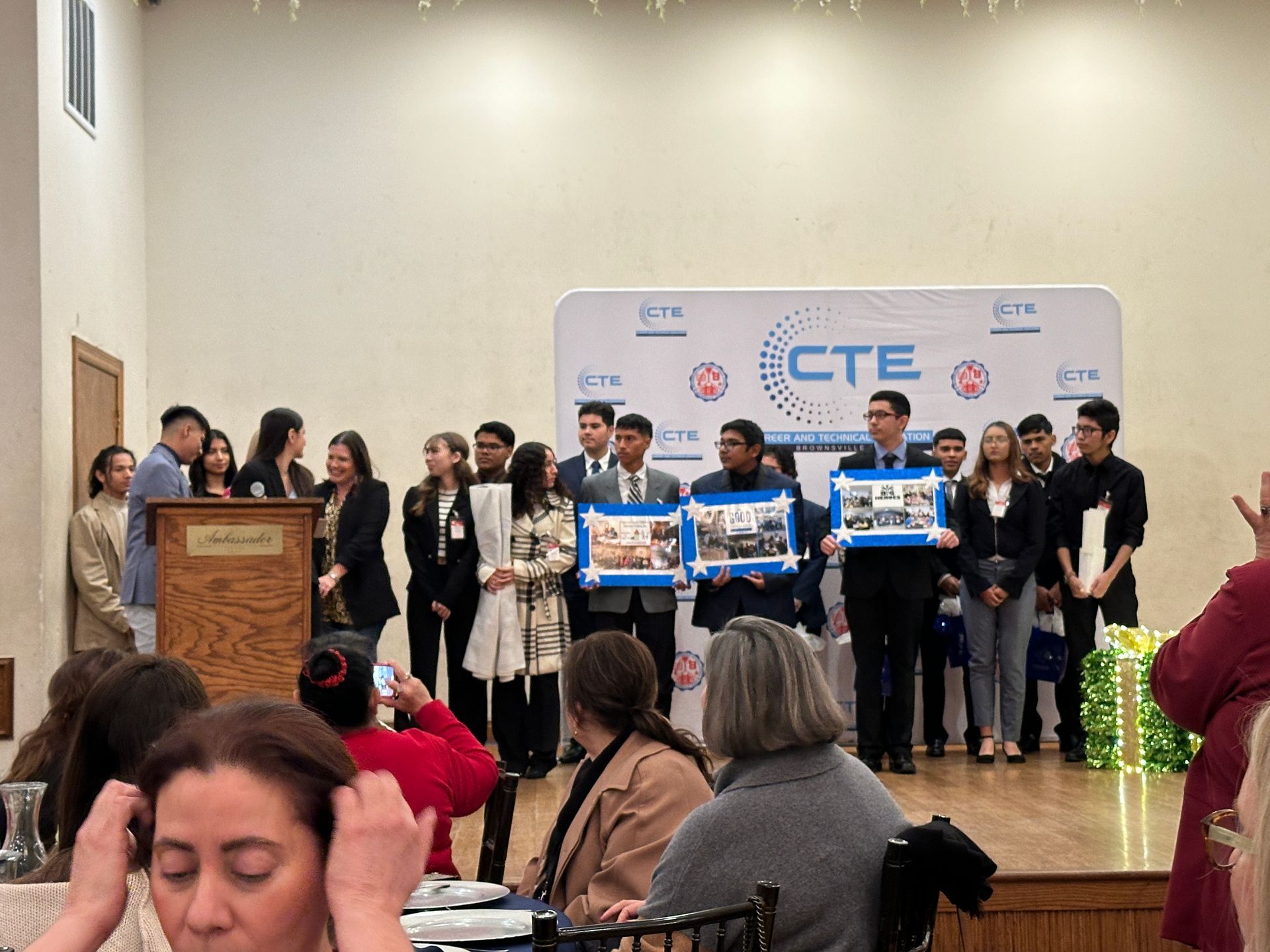 Students holding projects on stage at CTE event, with audience in front.