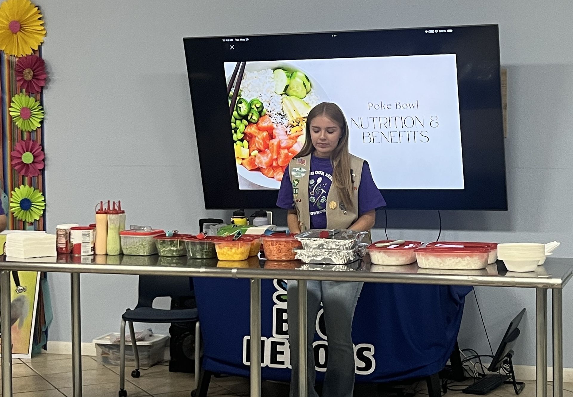 Girl Scout presenting a food demonstration with ingredients on a table. A screen behind her displays food.