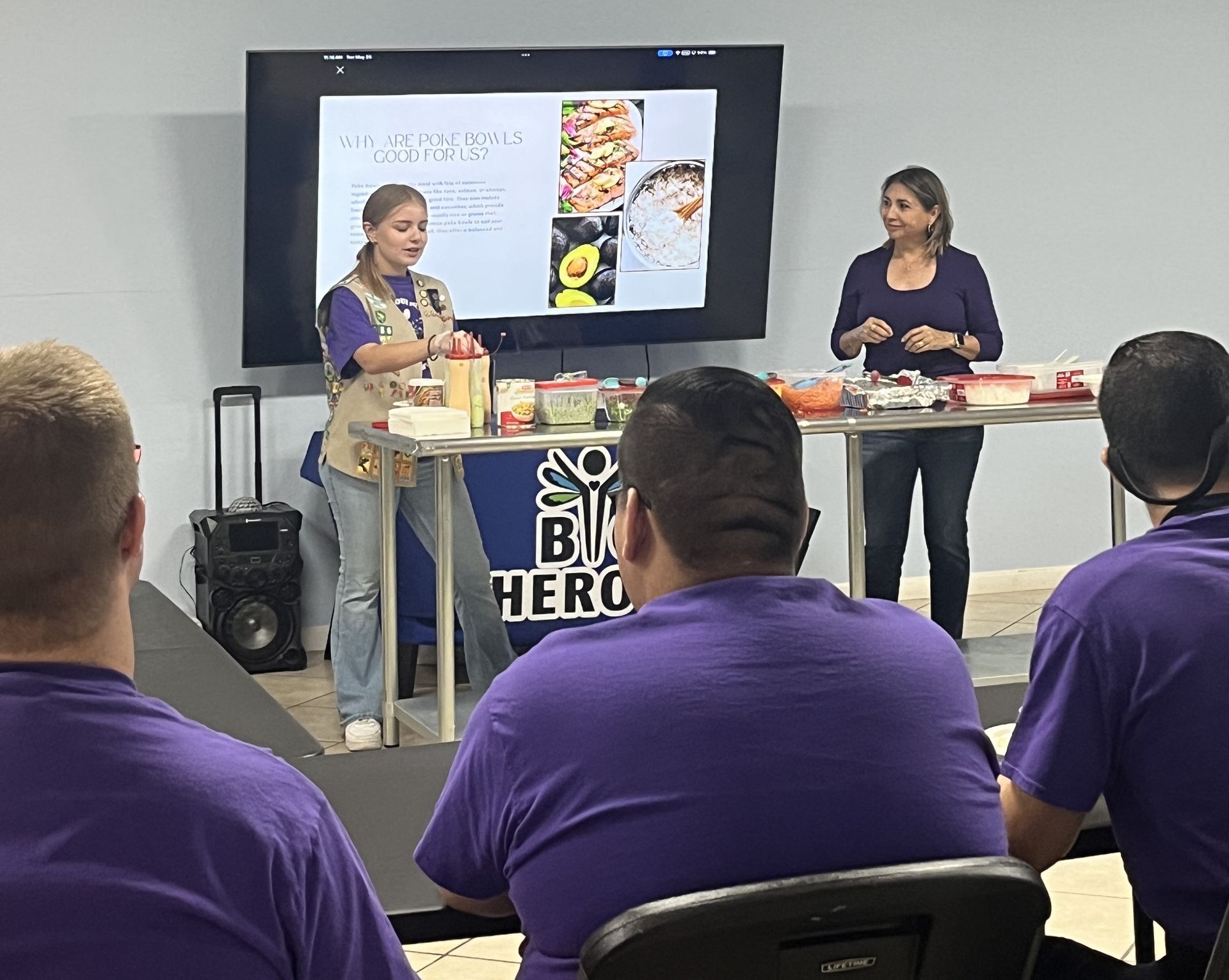 Two women give a cooking demonstration to a seated audience. Display screen shows food. People wear purple shirts.