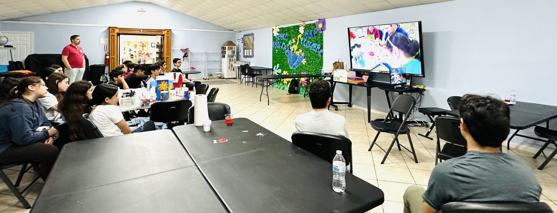 A group of people in a room watching a screen, some are seated at tables, while a man stands.