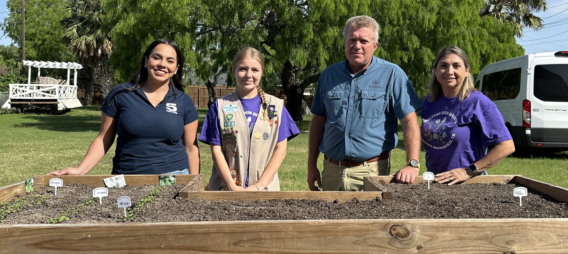Four people stand behind a raised garden bed. The group smiles at the camera. White van in the background.