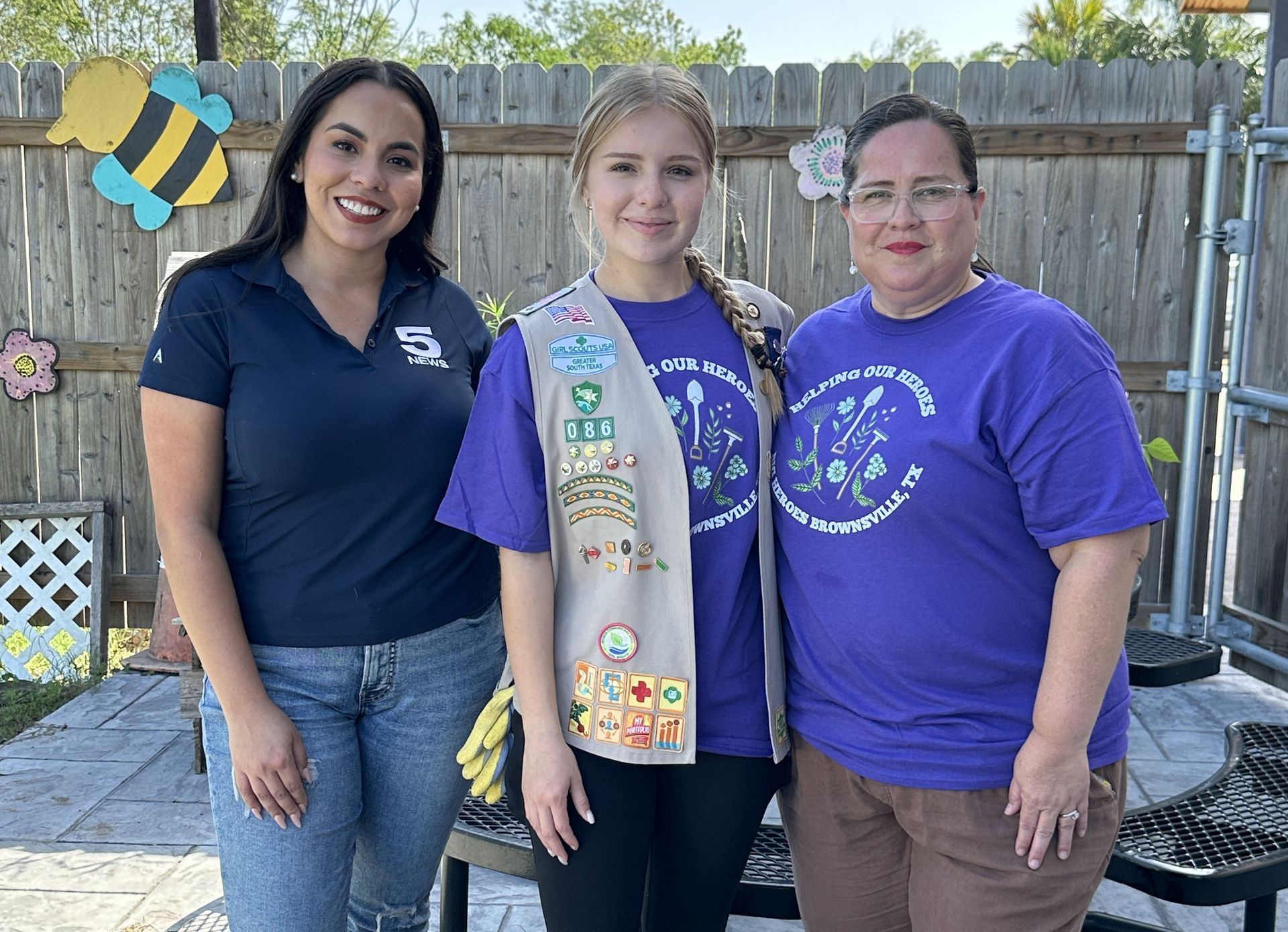 Three women smiling. A girl scout stands between two adults in a backyard setting.