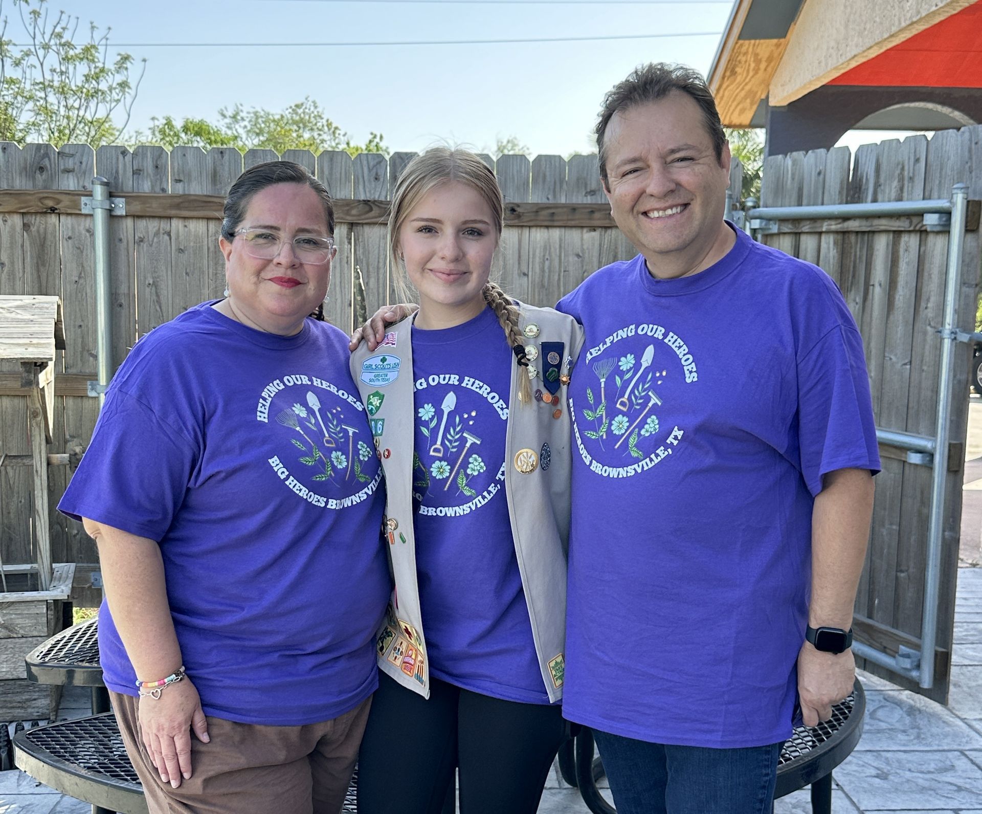 Three people in purple shirts pose in front of a fence; a woman, a young woman in a Girl Scout vest, and a man.