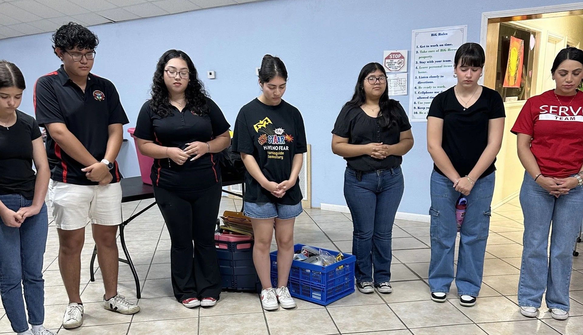 Group of people standing in a room, hands clasped. They appear to be in prayer.